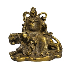 Op de afbeelding: Een messingkleurig standbeeld van een bebaarde krijger in traditioneel harnas die op een tijger zit. De figuur houdt een lange staf in één hand en is omringd door verspreide gouden munten en baren op een voetstuk met gedetailleerde reliëfs.