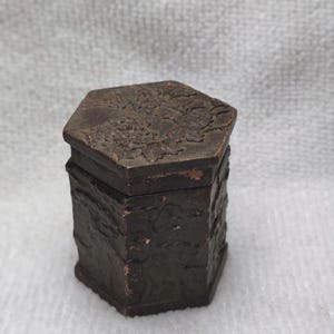 Pode incluir: Uma pequena caixa hexagonal marrom escuro com uma superfície texturizada. A tampa apresenta um design floral detalhado. A caixa tem uma aparência rústica e antiga, sugerindo que pode ser vintage ou artesanal.