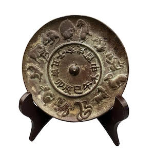 May include: A circular bronze-toned metal plate featuring a central raised knob surrounded by Chinese characters and a relief border of twelve zodiac animals. The plate has a weathered, dark patina and is displayed on a dark brown wooden stand.