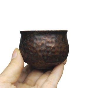 May include: A small, round, dark brown metal bowl featuring a distinct hammered texture across its exterior surface. The bowl has a slightly flared rim and a deep, rounded base, showcasing a weathered, metallic finish with subtle variations in color.