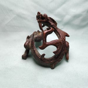 Puede incluir: Figura de dragón de bronce con detalles intrincados. El dragón está enroscado, con la cabeza levantada y la boca abierta. La escultura tiene una superficie texturizada y está sobre un fondo verde claro.