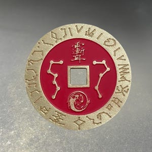 May include: A gold-coloured, circular coin with a red centre. The coin features embossed symbols and characters around the edge and in the centre. The centre has a square hole and a design of two intertwined fish.