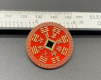 May include: A red and gold coin with Chinese characters and a square hole in the center. The coin is round and has a detailed design. The coin is approximately 1.5 inches in diameter.