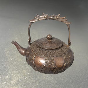 May include: Bronze teapot with a decorative handle and lid. The teapot features intricate embossed designs, including coins and a bat motif. The spout is curved, and the handle is arched. The teapot is set against a gray background.