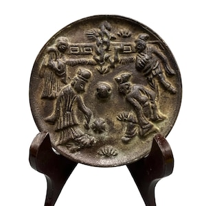 Op de afbeelding: Een ronde, bronskleurige metalen spiegel met een reliëf van vier figuren in traditionele kleding rondom een centrale knop. Het oppervlak toont ingewikkelde patronen van gebladerte en gestileerde motieven met een donkere, verweerde afwerking.