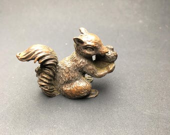 May include: A small, bronze-colored squirrel figurine holding a nut. The squirrel is in a seated position with its bushy tail curled up. The figurine has detailed fur and facial features, and is set against a dark background.