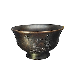 Puede incluir: Un cuenco de metal en tono bronce oscuro con un borde ancho y acampanado y una base circular elevada. La superficie exterior está decorada con patrones en relieve que representan nubes estilizadas y caracteres caligráficos tradicionales.