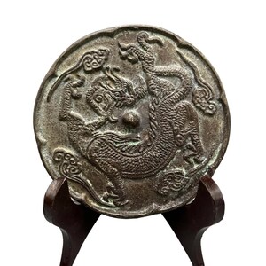 Op de afbeelding: Een ronde, bronskleurige metalen spiegel met een gedetailleerd reliëf van een draak met schubben en klauwen, omringd door gestileerde wolkenmotieven. Het oppervlak heeft een verweerde, donkere patina met sporen van groene oxidatie.