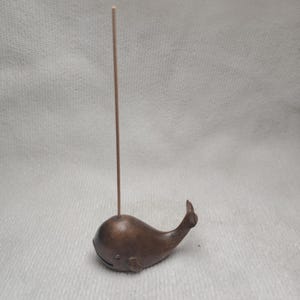 May include: A bronze-colored whale-shaped incense holder with a slender, upright incense stick. The whale has a rounded body, a small fin, and a smiling face. The holder is designed to hold incense sticks.