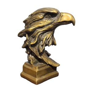 May include: A bronze-toned sculpture of an eagle head in profile, featuring detailed feathers and a sharp, curved beak. The eagle head is mounted on a square, tiered base, showcasing a metallic finish with dark highlights in the crevices.
