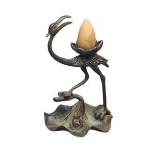 Puede incluir: Porta incienso de bronce con forma de grulla, sosteniendo un incienso en forma de cono. La grulla se apoya en una hoja de nenúfar con una pequeña rana. El soporte mide aproximadamente 15 cm de altura.
