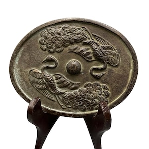 Puede incluir: Un espejo ovalado de metal en tono bronce que presenta un diseño en relieve de dos pavos reales con colas desplegadas frente a frente. Un pequeño botón circular elevado está situado en el centro del espejo, rodeado por las detalladas figuras aviares.