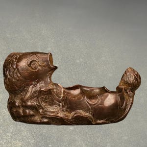 May include: A bronze-colored, fish-shaped decorative object. The sculpture features a detailed fish head with an open mouth and a curved body. The tail end is also detailed. The object appears to be a small container or holder.