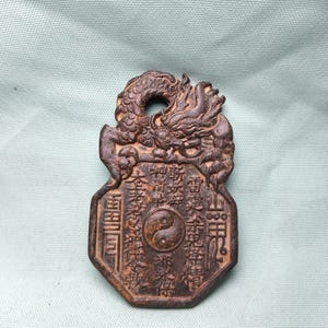 May include: A bronze-colored, rectangular pendant with a dragon design at the top and a hole for hanging. The pendant features intricate carvings, including Chinese characters and a yin-yang symbol. The overall design is detailed and ornate.