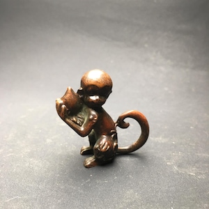 May include: A small, bronze-colored monkey figurine holding a small object. The monkey is in a seated position with its tail curled. The figurine has detailed features and a smooth finish. The background is a solid gray.