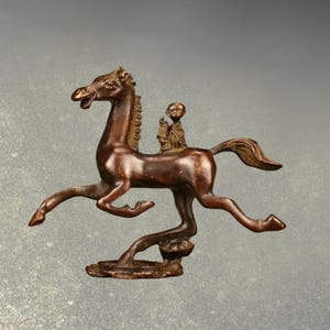 May include: A bronze sculpture of a horse with a rider. The horse is in mid-stride, with its legs extended. The rider is small and seated on the horse's back. The sculpture has a dark brown patina and is set on a small base.