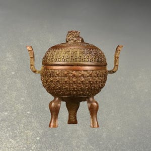 May include: A bronze-coloured incense burner with a round body and a lid. The lid is topped with a small decorative figure. The burner has three legs and two handles. The body and lid are covered in a detailed pattern.