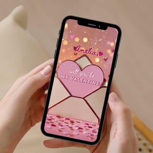 May include: A smartphone displays a Valentine's Day themed graphic. The screen shows a pink heart-shaped envelope with the text "Amelia, will you be my Valentine?" surrounded by pink confetti and hearts. The phone is held by hands.