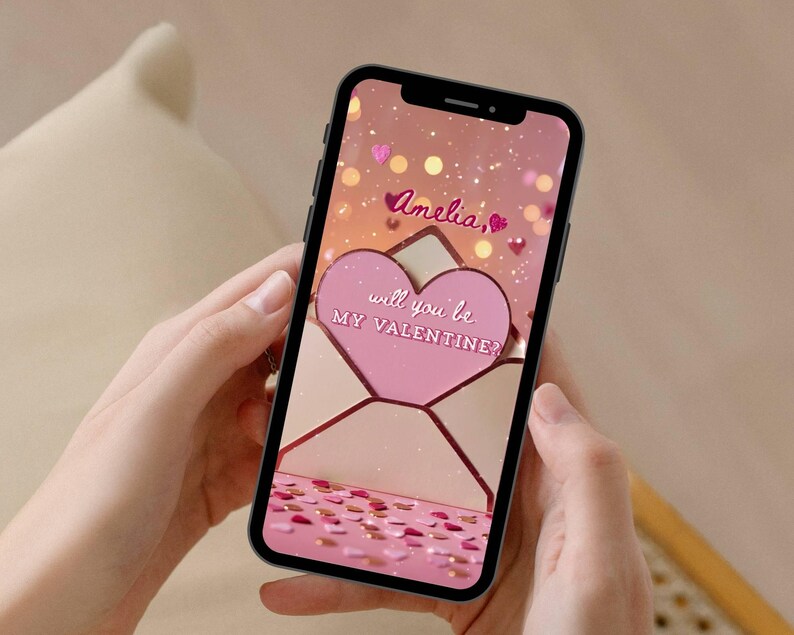 May include: A smartphone displays a Valentine's Day themed graphic. The screen shows a pink heart-shaped envelope with the text "Amelia, will you be my Valentine?" surrounded by pink confetti and hearts. The phone is held by hands.