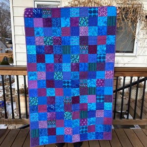 May include: A purple and blue patchwork quilt with a variety of patterns. The quilt is made up of squares of different fabrics, each with a unique design. The quilt is being held up by a person, and the bottom of the quilt is resting on a wooden floor.
