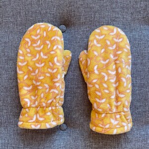 May include: A pair of yellow fleece mittens with a white floral pattern. The mittens have a gathered cuff.