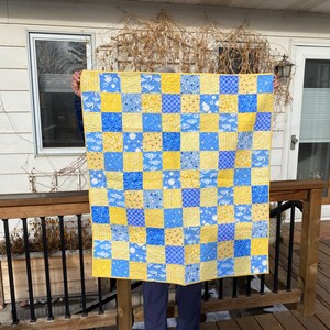 May include: A blue and yellow patchwork quilt with a checkered pattern. The quilt is held up by a person in front of a wooden deck.