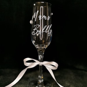 May include: A clear champagne flute with the words "Mums Bubbly" in white script. The glass is decorated with small white dots and a white satin ribbon tied around the stem. The background is dark.