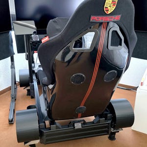 Könnte beinhalten: Schwarzer Rennsimulator-Sitz mit Porsche-Logo und roten Akzenten. Der Sitz hat einen schwarzen Rahmen und ist für ein realistisches Fahrerlebnis konzipiert. Der Sitz hat eine hohe Rückenlehne und ein komfortables Design.