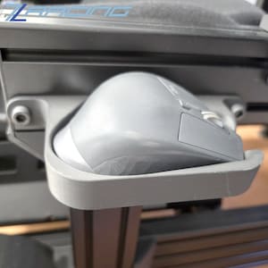 Könnte beinhalten: Eine graue Computermaus liegt in einer maßgefertigten, grauen Halterung. Die Halterung ist an einem schwarzen Rahmen befestigt, wahrscheinlich Teil eines Rennsimulator-Setups. Die Maus hat ein Scrollrad und mehrere Tasten.