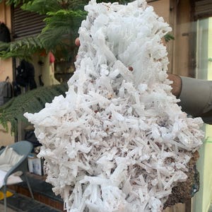 Puede incluir: Un gran grupo de cristales blancos con una superficie rugosa y texturizada. Los cristales están densamente agrupados, dando una apariencia puntiaguda. La base del grupo es una mezcla de roca marrón y marrón rojizo.