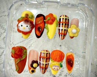 May include: A set of ten artificial nails in a clear plastic case. The nails feature various designs, including a Hello Kitty face, a flower, a pumpkin, and a plaid pattern. Colors include orange, yellow, brown, and pink.