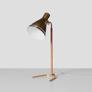 May include: A brass desk lamp with a conical shade and a light brown wooden stem. The lamp has a three-legged base and a white interior shade. The lamp is plugged into a white electrical outlet.
