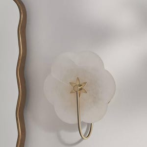 May include: A white, flower-shaped wall sconce with a gold-colored metal stem and hook. The sconce is mounted on a white wall, and a gold-framed mirror is partially visible on the left. The sconce has a star-shaped detail.
