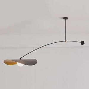 May include: Modern black and gold pendant light fixture. The fixture has a curved arm with a circular light at the end and a flat, petal-shaped shade with a light bulb. The light is suspended from the ceiling.