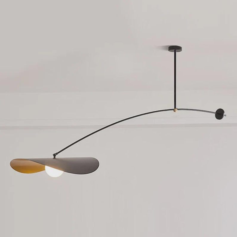 May include: Modern black and gold pendant light fixture. The fixture has a curved arm with a circular light at the end and a flat, petal-shaped shade with a light bulb. The light is suspended from the ceiling.