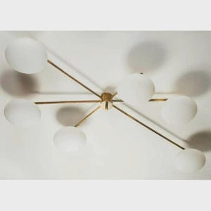 Peut inclure: Un plafonnier moderne avec six abat-jours globulaires blancs. Le luminaire a une finition en laiton et est monté sur un plafond blanc. Les abat-jours sont espacés uniformément autour d'un moyeu central.