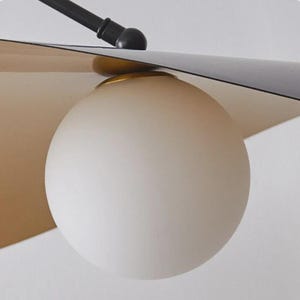 May include: Close-up of a modern light fixture featuring a frosted white globe light. The fixture has a black arm and a gold and tan shade. The design is minimalist and contemporary, suitable for home decor.