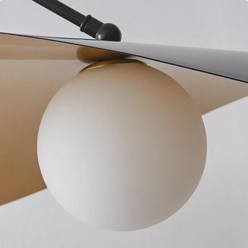 May include: Close-up of a modern light fixture featuring a frosted white globe light. The fixture has a black arm and a gold and tan shade. The design is minimalist and contemporary, suitable for home decor.
