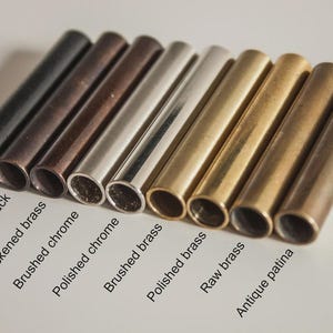 May include: A collection of nine cylindrical metal tubes arranged in a row, featuring various finishes labeled as matte black, antique black, blackened brass, brushed chrome, polished chrome, brushed brass, polished brass, raw brass, and antique patina.