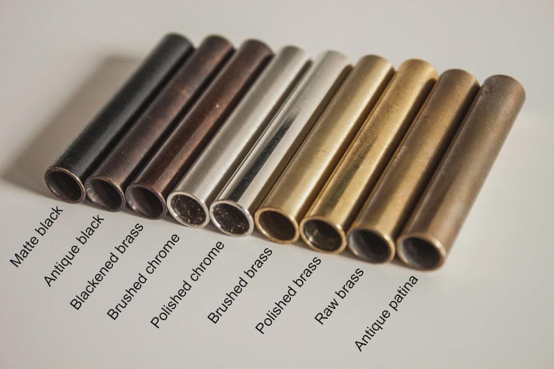 May include: A collection of nine cylindrical metal tubes arranged in a row, featuring various finishes labeled as matte black, antique black, blackened brass, brushed chrome, polished chrome, brushed brass, polished brass, raw brass, and antique patina.