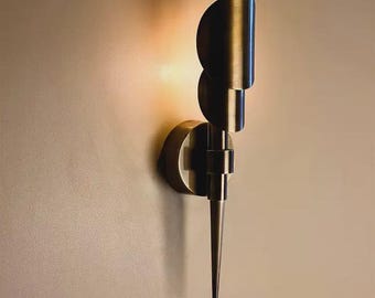 May include: A modern wall sconce with a brushed metal finish. The sconce features a semi-circular shade and a long, pointed base. The light is warm and creates a soft glow on the wall. The design is minimalist and contemporary.