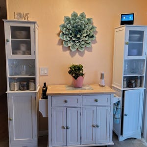 May include: A kitchen scene featuring white cabinets, a wooden countertop, and a central island. A large, decorative, green flower hangs on the wall. Glassware and jars are visible inside the cabinets. A small plant in a pink pot sits on the countertop.