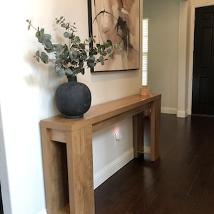 May include: A light oak wood console table with a black vase holding green leafy branches. A framed abstract painting hangs above the table. The table is 60 inches long.