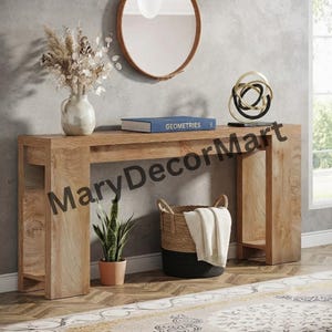May include: A light brown wooden console table with a rectangular top and two side supports. On the table sits a white vase with dried flowers, a blue book titled GEOMETRIES, and a black and gold abstract sculpture. A basket sits below.