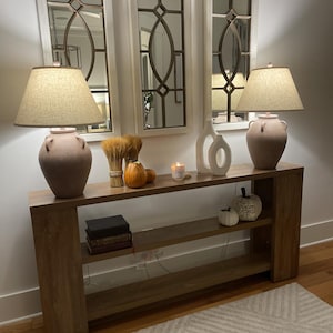 May include: A wooden console table with two lamps, decorative mirrors, and fall-themed decor. The lamps have beige shades and sit on pink ceramic bases. The table is adorned with pumpkins, wheat stalks, and a candle, creating a warm, inviting atmosphere.