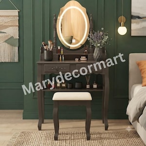 May include: A dark gray vanity table with an oval LED-lit mirror, two drawers, and a lower shelf. The set includes a matching stool with a cream-colored upholstered seat. Various cosmetic bottles and makeup brushes are arranged on the surface.