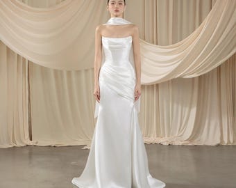 May include: A strapless white satin gown with a draped bodice and a flowing skirt. The dress features a matching scarf draped over the shoulders. The background includes beige fabric curtains, creating a soft, elegant aesthetic.