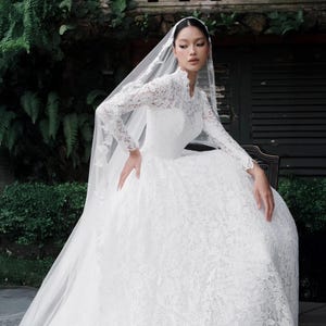 Può includere: Un abito da sposa in pizzo bianco con maniche lunghe e collo alto. L'abito presenta una gonna ampia ed è abbinato a un lungo velo trasparente. La sposa è seduta, con le mani appoggiate sulle ginocchia.