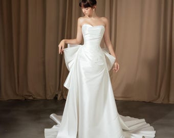 May include: A strapless white wedding gown with a large bow detail at the back. The dress features a fitted bodice and a flowing skirt that extends into a train. The fabric appears to be a smooth, satin-like material.