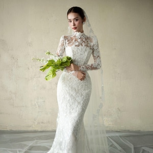 Puede incluir: Vestido de novia de encaje blanco con mangas largas y cuello alto. El vestido tiene un corpi&ntilde;o ajustado y una falda de sirena. Un velo transparente y un ramo de flores blancas y verdes completan el look nupcial.